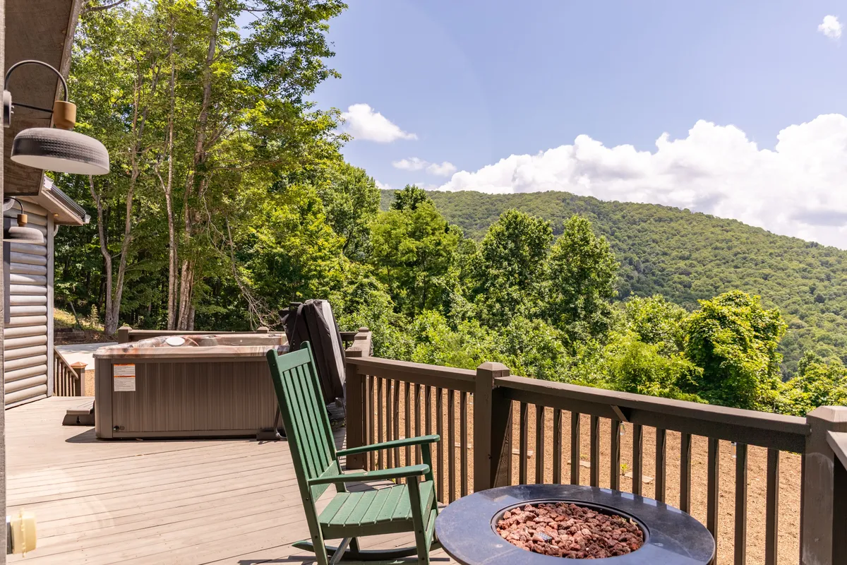 An inviting exterior scene showing a green Adirondack chair near a fire pit, with the hot tub just behind. The area is set against a sweeping mountain view, creating a cozy spot for gathering around the fire or relaxing in the tub, all with a panoramic natural backdrop.