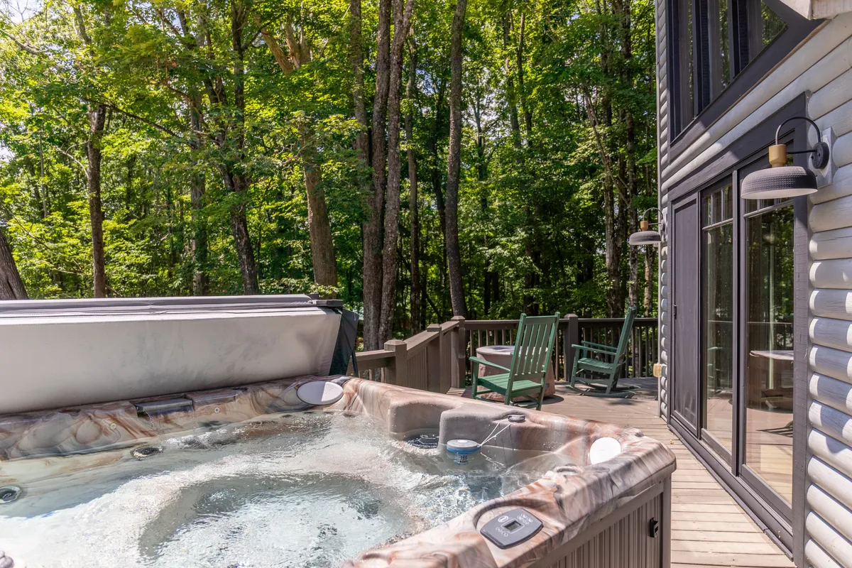 This image features a serene outdoor deck area with a private hot tub, surrounded by lush trees. There are deck chairs positioned for relaxation, inviting guests to enjoy the natural mountain setting. The atmosphere is tranquil, perfect for unwinding, socializing, or stargazing from the comfort of the hot tub.