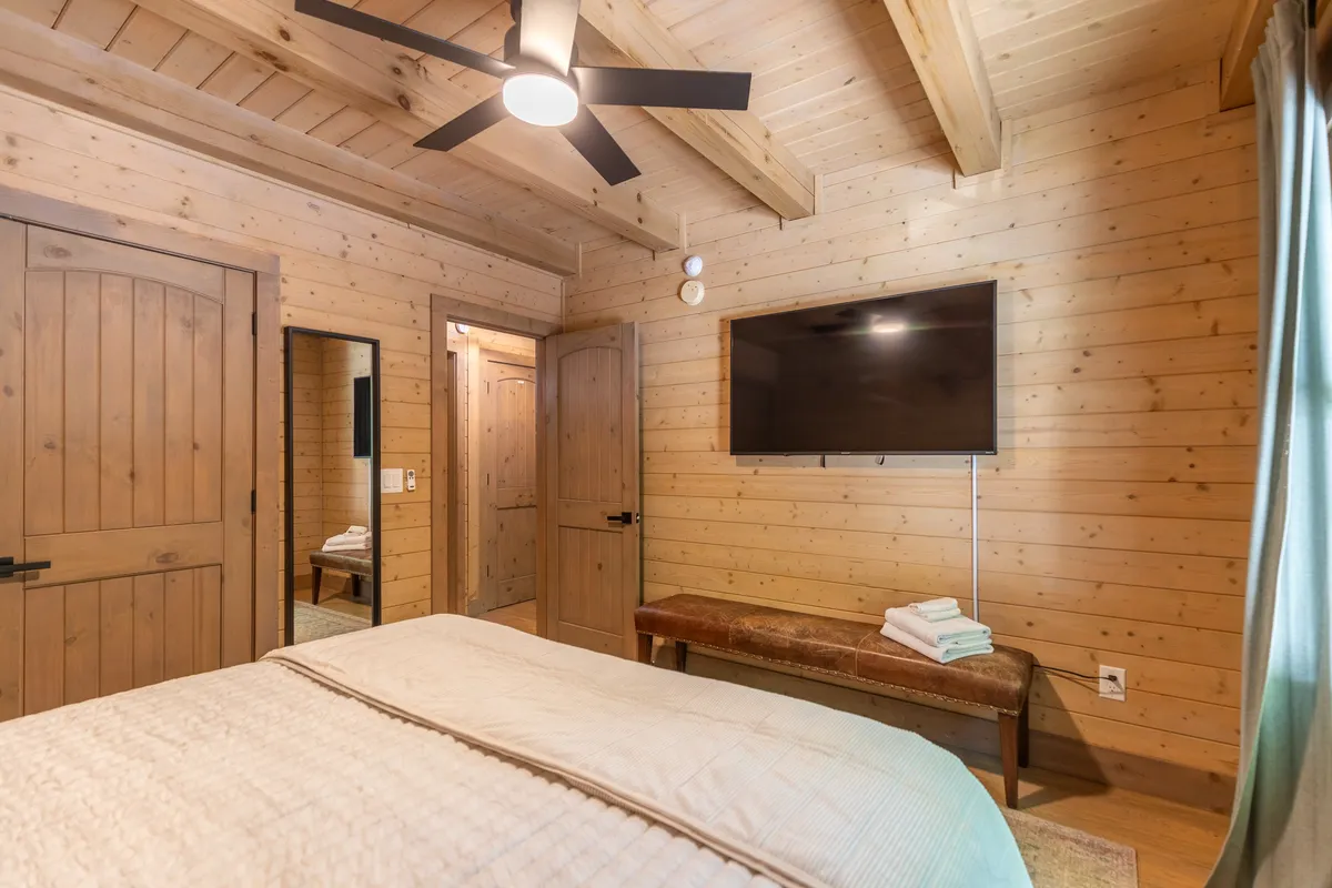 A calm and comfortable bedroom with textured bedding, wooden ceiling beams, and a television mounted on the wall. A mirror and wooden doors complete the space, suggesting an ensuite arrangement.