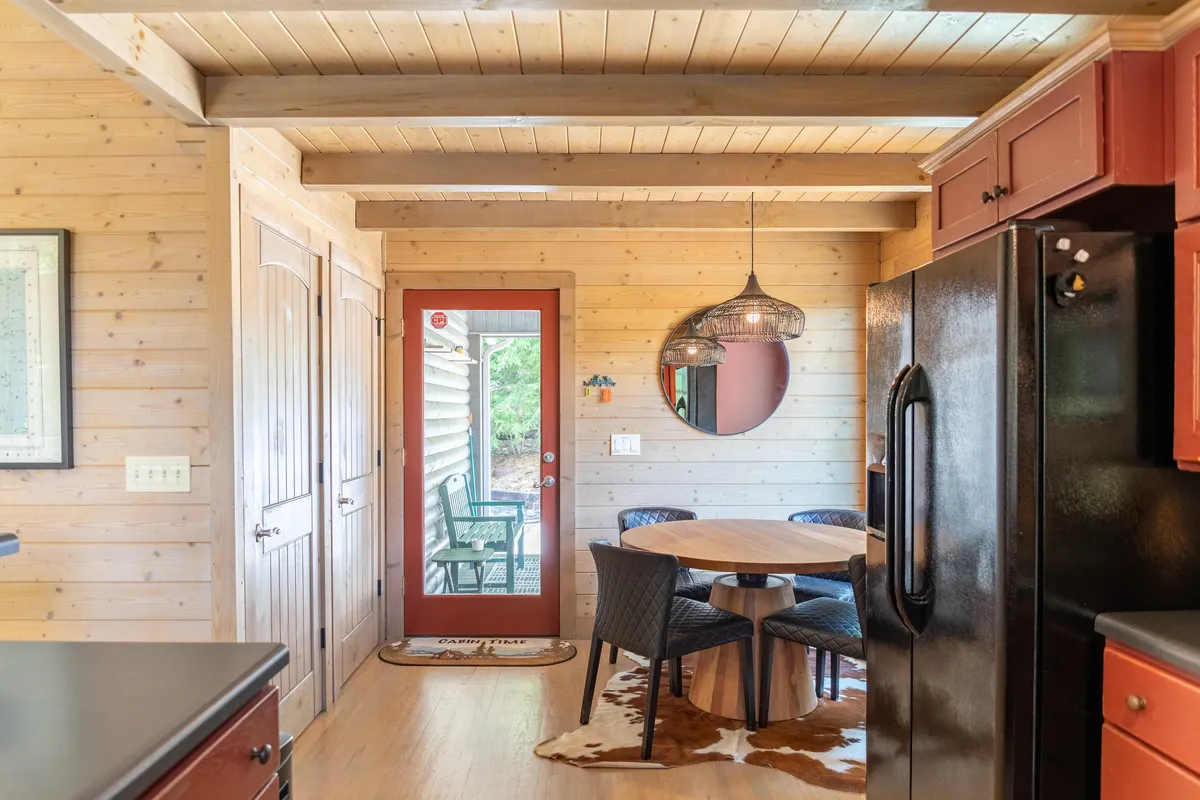 A round dining table sits beneath a hanging light fixture, with a red door leading directly outside. The cozy setup invites intimate family meals or morning coffee, while the easy access to the outdoors alludes to deck gatherings and sunset cocktails.
