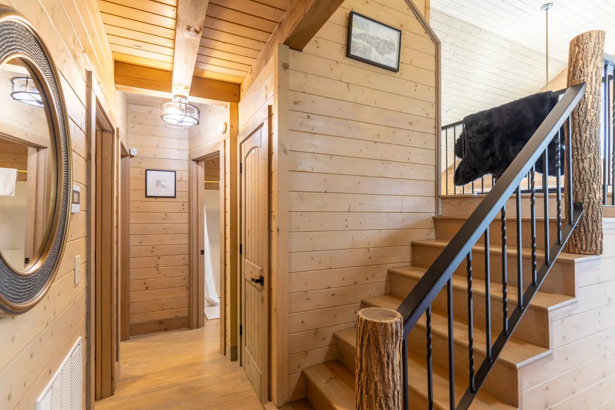 The image captures the cabin's entryway, featuring a wooden staircase, round mirror, and rustic decor. Natural light filters through the space, highlighting the inviting textures and the warm, welcoming atmosphere as guests enter the cabin.