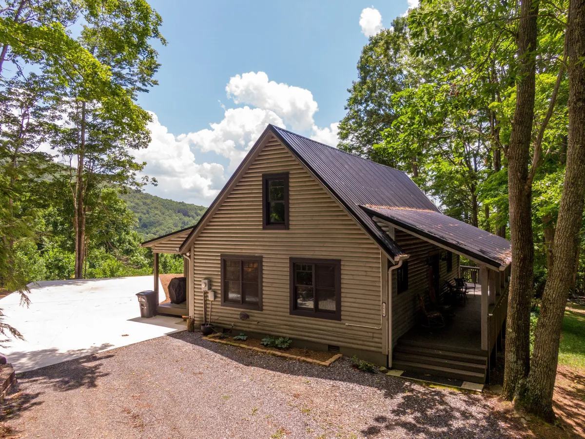 This exterior shot features the Queen Ridge Cabin nestled among tall trees, with a peaceful driveway leading up to the house. The generous porch is visible, set against a tranquil natural backdrop, evoking a sense of privacy and immersion in nature.