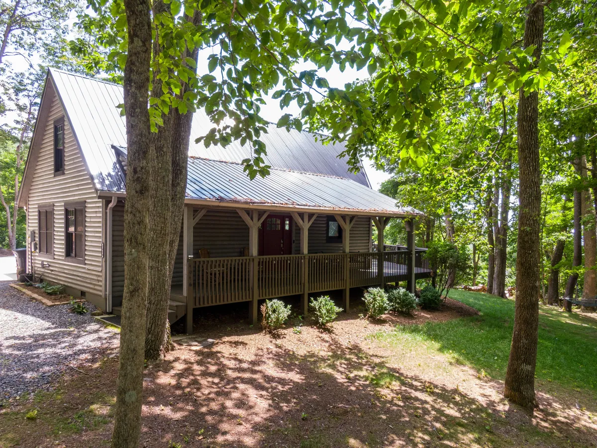 This image showcases the inviting front porch of Queen Ridge Cabin, complete with a red door, wooden railing, and surrounded by lush trees. The space is set up for outdoor relaxation and socializing, hinting at the welcoming atmosphere awaiting guests.