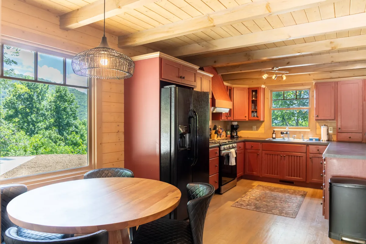 A view of the cabin's kitchen, showcasing red cabinets, a black refrigerator, a wooden table, and a kitchen island. Large windows bring in plenty of natural light and offer views of the outdoors, connecting the kitchen to the surrounding scenery.