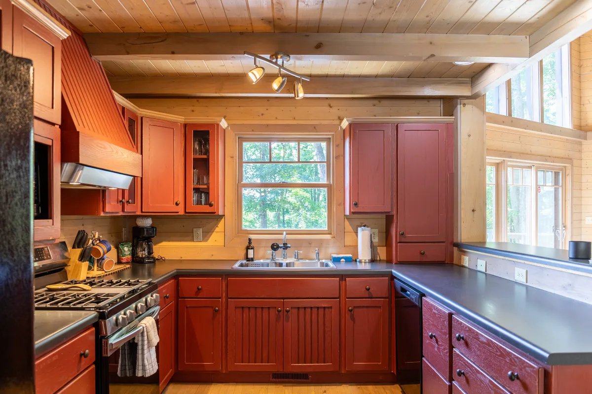 A warm kitchen scene with rich red cabinetry, stainless steel appliances, and a window above the sink. The kitchen is set up for group cooking and family gatherings, with ample counter space and a welcoming atmosphere.