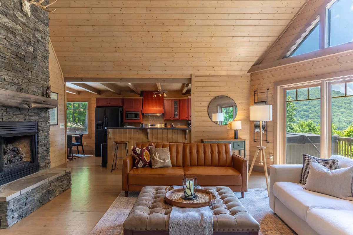 This image presents a warm, open-concept living space featuring a stone fireplace, leather sofa, and inviting kitchen area. Large windows in the background bathe the room in natural light and offer glimpses of the outdoors, while the kitchen’s proximity to the living room highlights the fluidity between spaces.