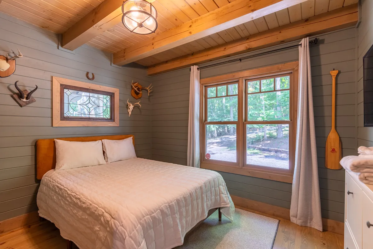This image features a peaceful bedroom with a neatly made bed, a window offering views of the surrounding nature, and decorative wall art that enhances the tranquil atmosphere. The natural light streaming in suggests a restful and airy environment.