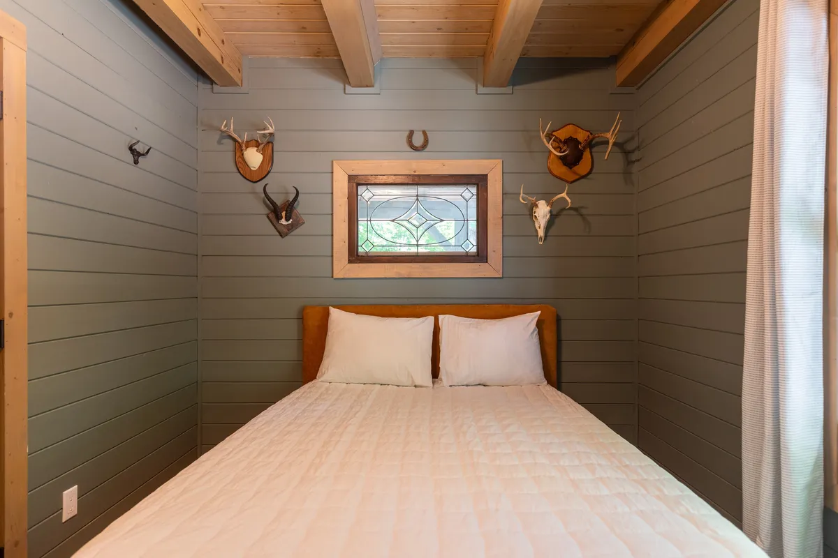 A cozy bedroom with a bed set against a backdrop of a stained glass window and animal head mounts. Warm light fills the room, and the decor evokes a sense of comfort and rustic charm.