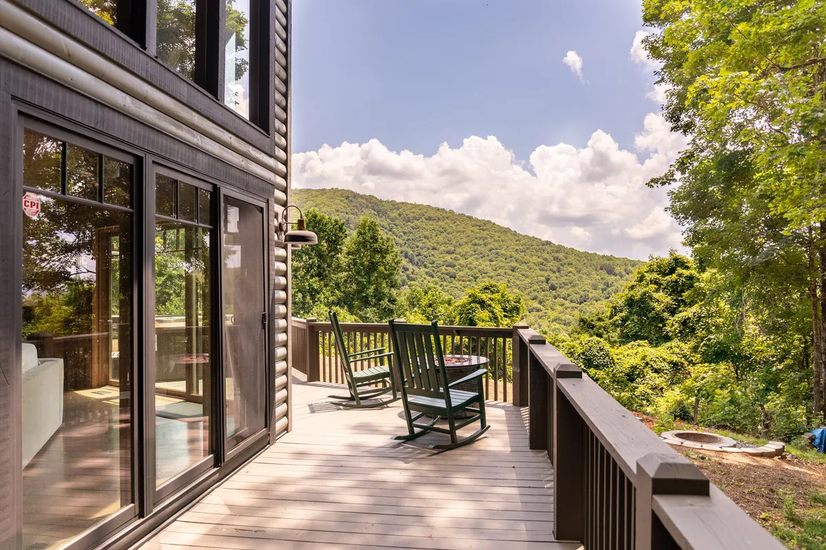 An inviting wraparound deck set against panoramic mountain backdrops, equipped with rocking chairs for two. The deck's vantage captures the quiet beauty of sunrise and offers a serene spot for morning coffee or evening stargazing, nestled in lush greenery.